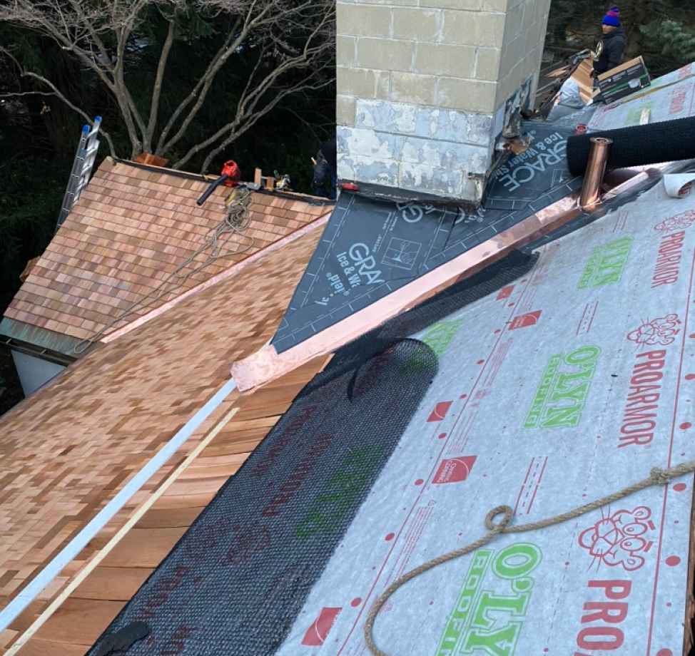 Cedar shingle roof installation in progress with synthetic underlayment and copper flashing near chimney.