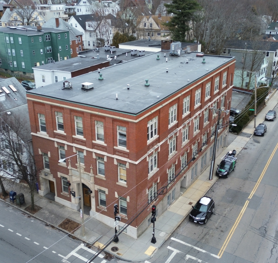 Aerial view of a large red brick apartment building with a flat black rubber roof in an urban neighborhood.