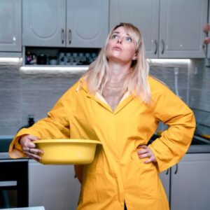 A frustrated homeowner in a yellow raincoat holding a bucket under a roof leak in the kitchen.