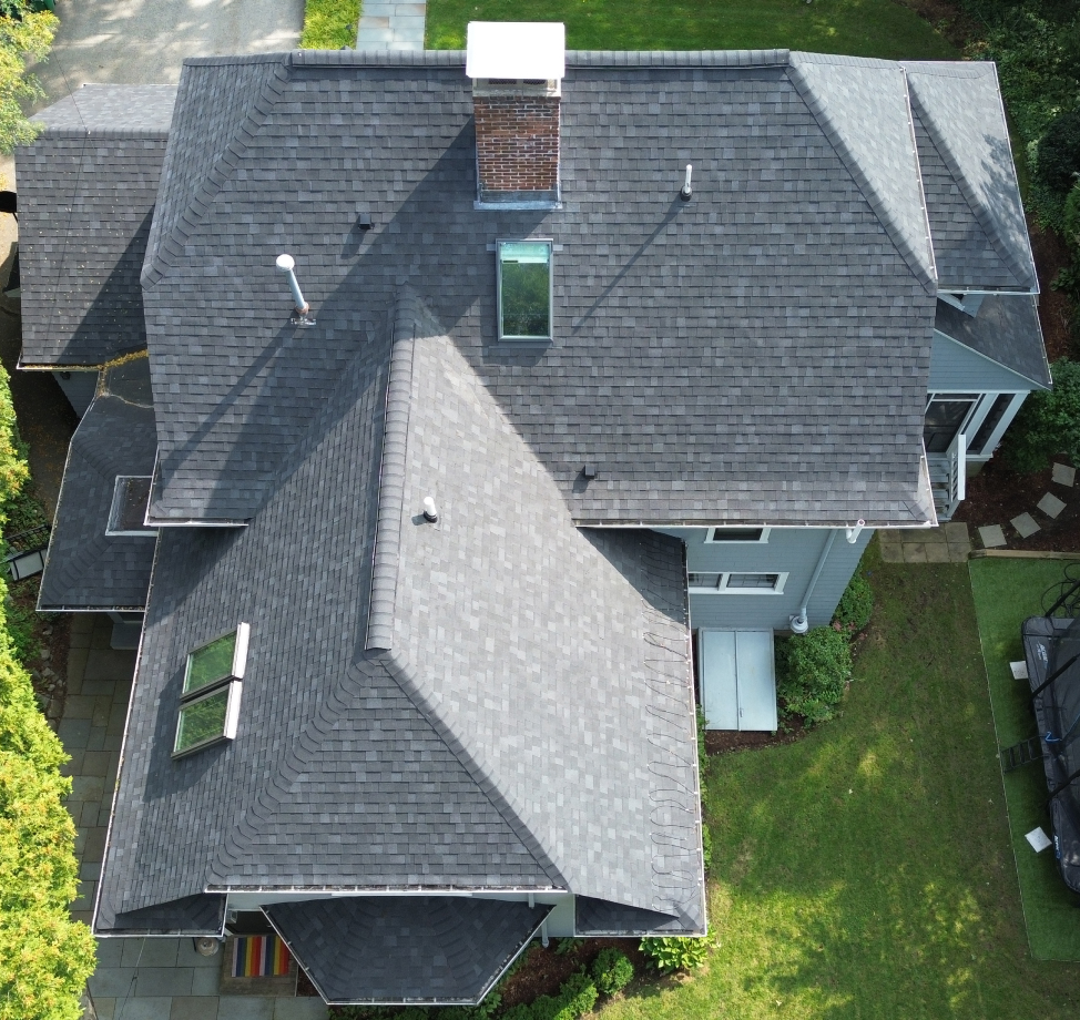 Aerial view of a gray shingle roof with multiple pitches, skylights, and a central brick chimney.