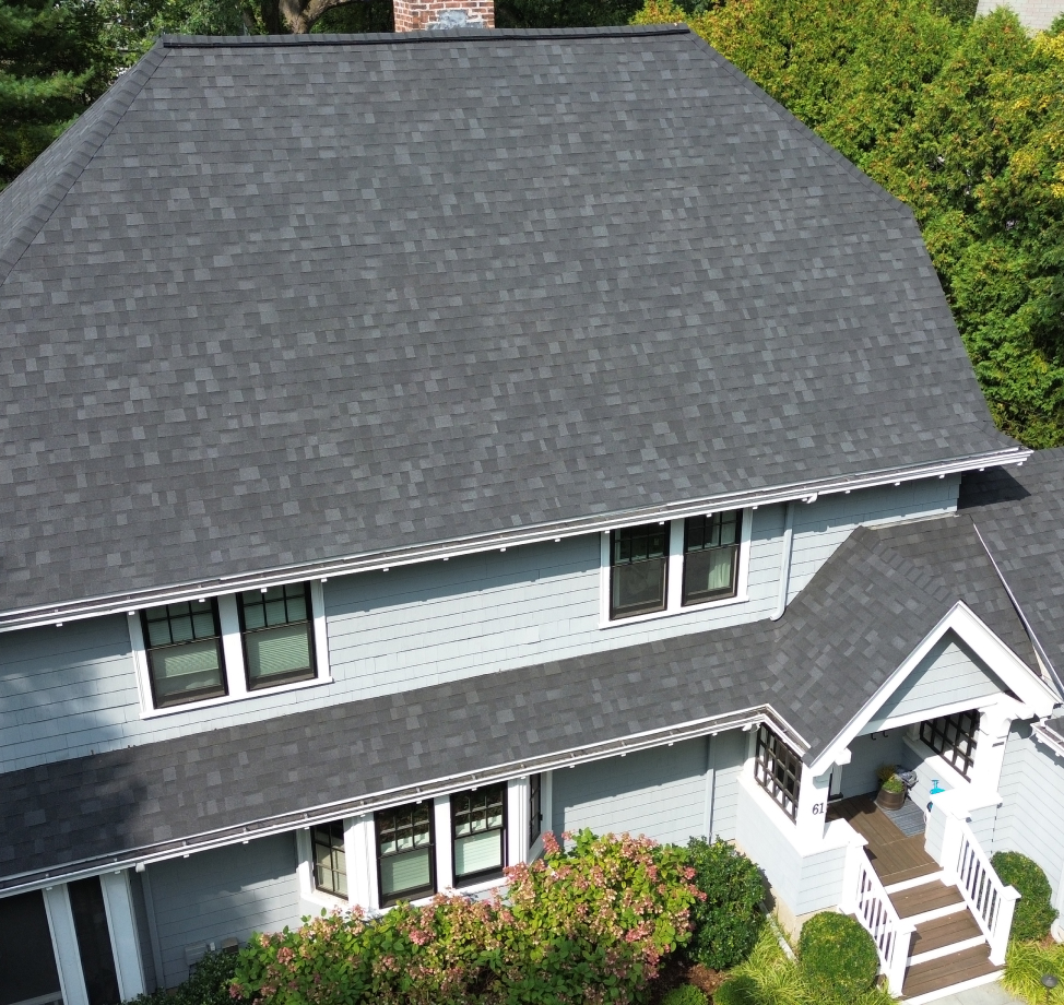 Aerial view of a light blue house with new dark gray asphalt shingle roof, white trim, and lush landscaping.