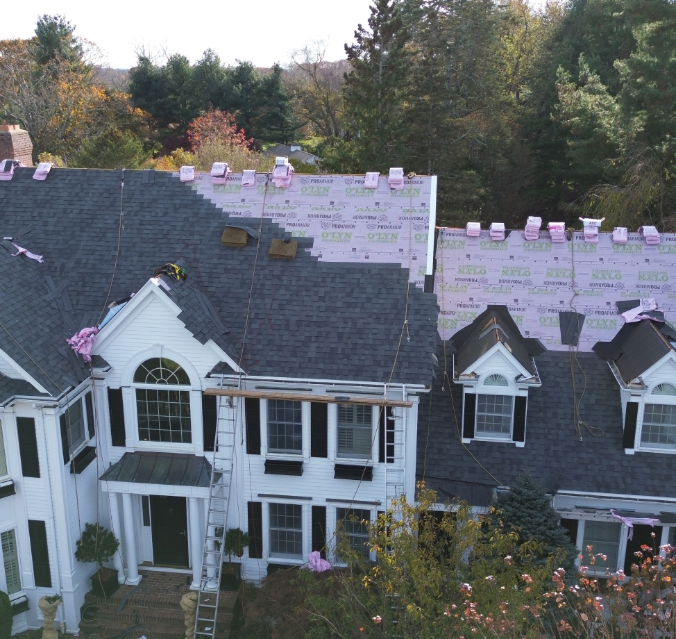 Large white colonial home mid-roof replacement with shingles partially installed and pink underlayment visible.