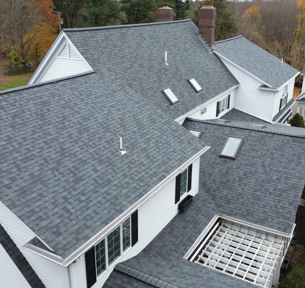 Complex roofline with gray asphalt shingles, white siding, skylights, and brick chimneys in autumn setting.